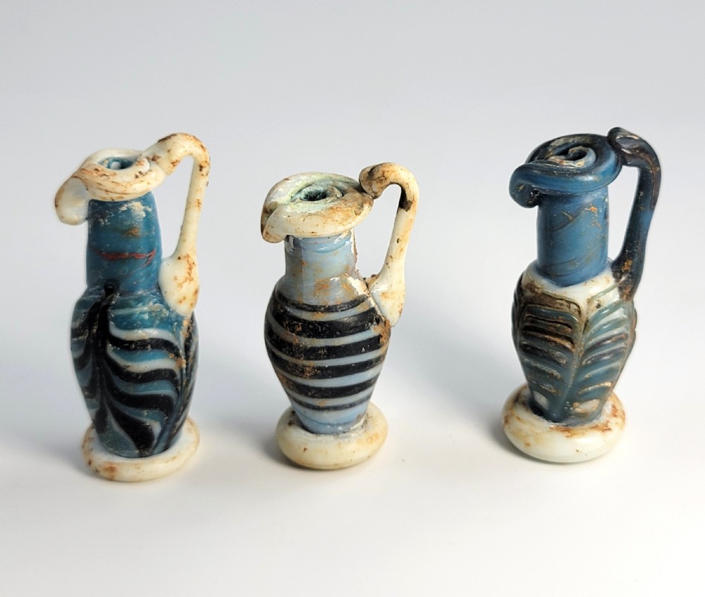 A set of four small ancient Roman glass unguentaria with blue bodies, white trailed decoration, and small handles resting inside a cardboard box. View V