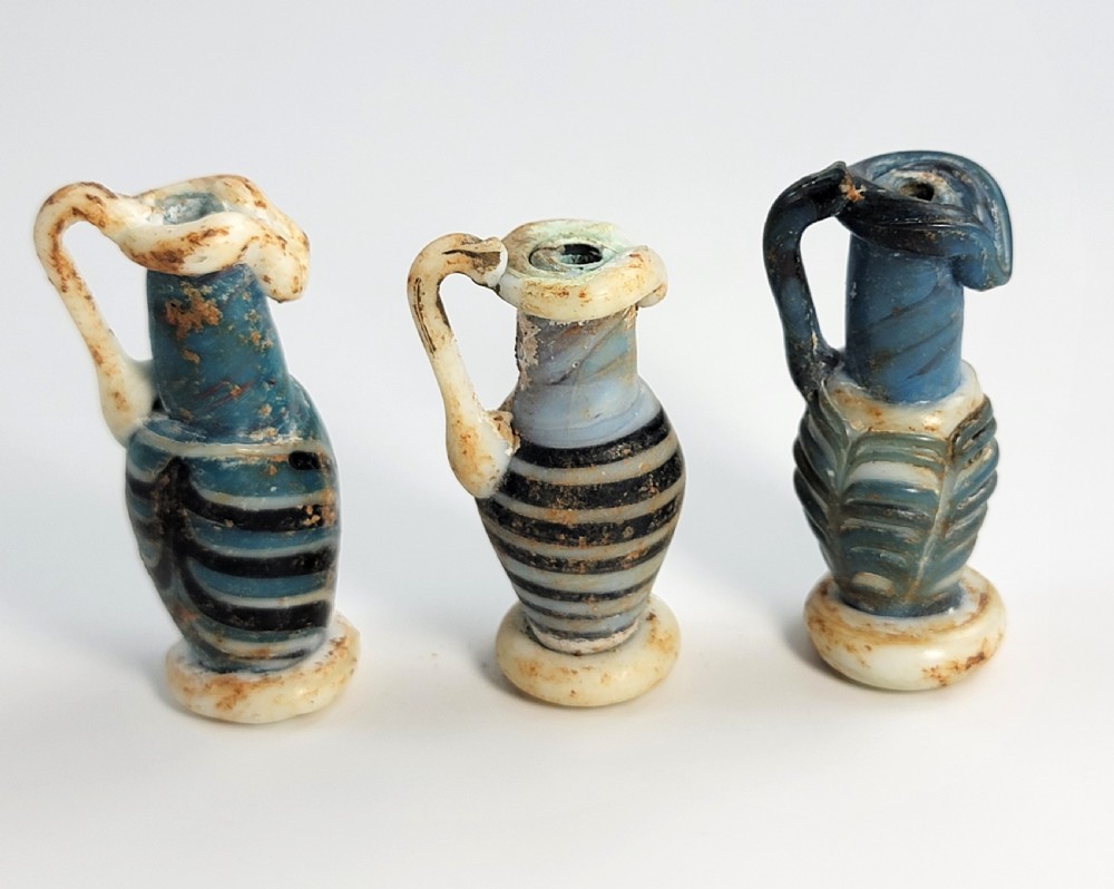 A set of four small ancient Roman glass unguentaria with blue bodies, white trailed decoration, and small handles resting inside a cardboard box. View VII