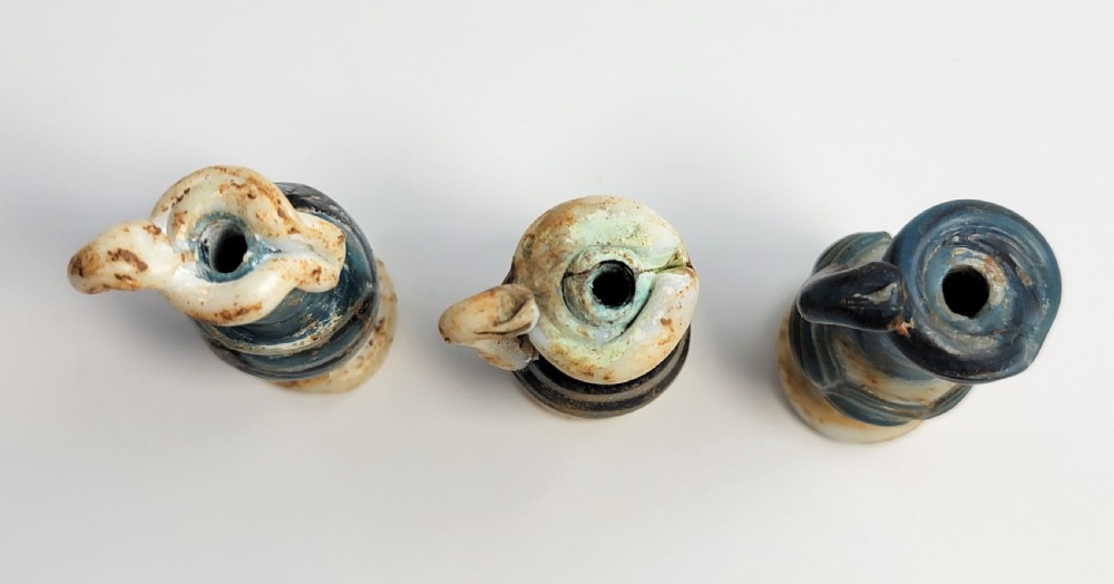 A set of four small ancient Roman glass unguentaria with blue bodies, white trailed decoration, and small handles resting inside a cardboard box. View VIII