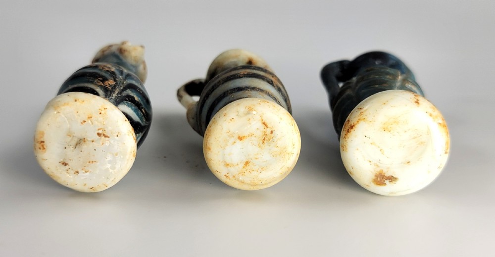 A set of four small ancient Roman glass unguentaria with blue bodies, white trailed decoration, and small handles resting inside a cardboard box. View IX