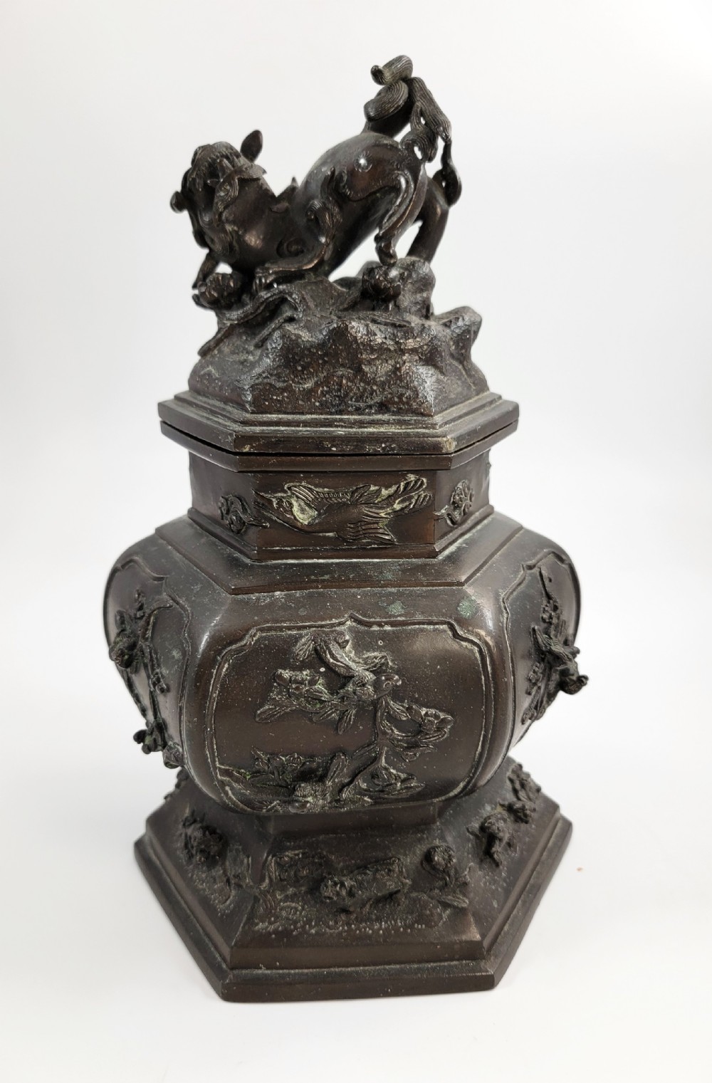 A dark patinated hexagonal bronze incense burner featuring floral relief panels and topped with a sculpted Fu lion finial.