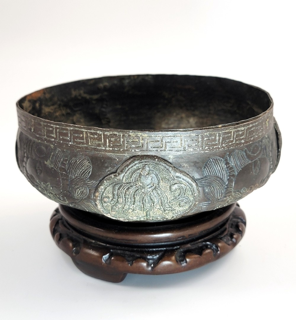 A patinated dark bronze Japanese bowl with carved archaic relief patterns and a key-fret border, resting on a carved wooden stand.