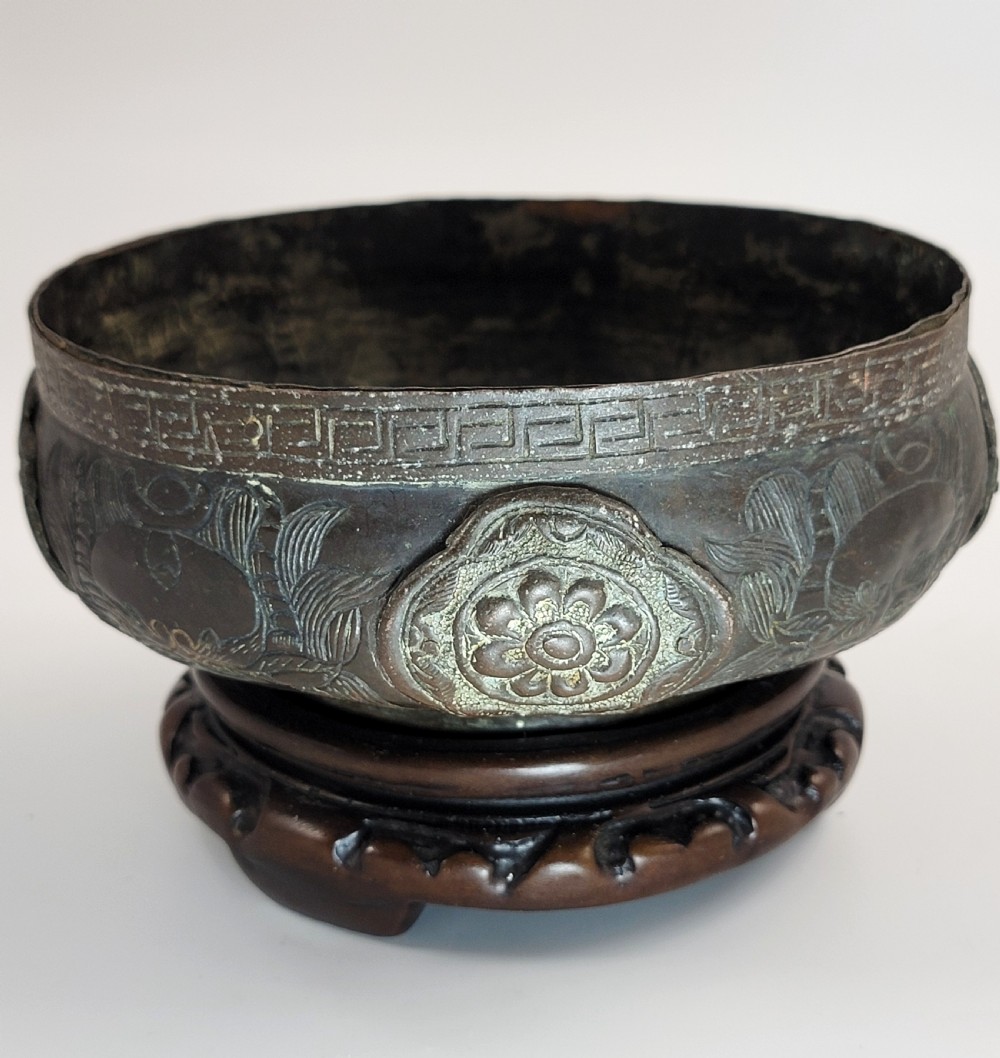 A patinated dark bronze Japanese bowl with carved archaic relief patterns and a key-fret border, resting on a carved wooden stand. View III