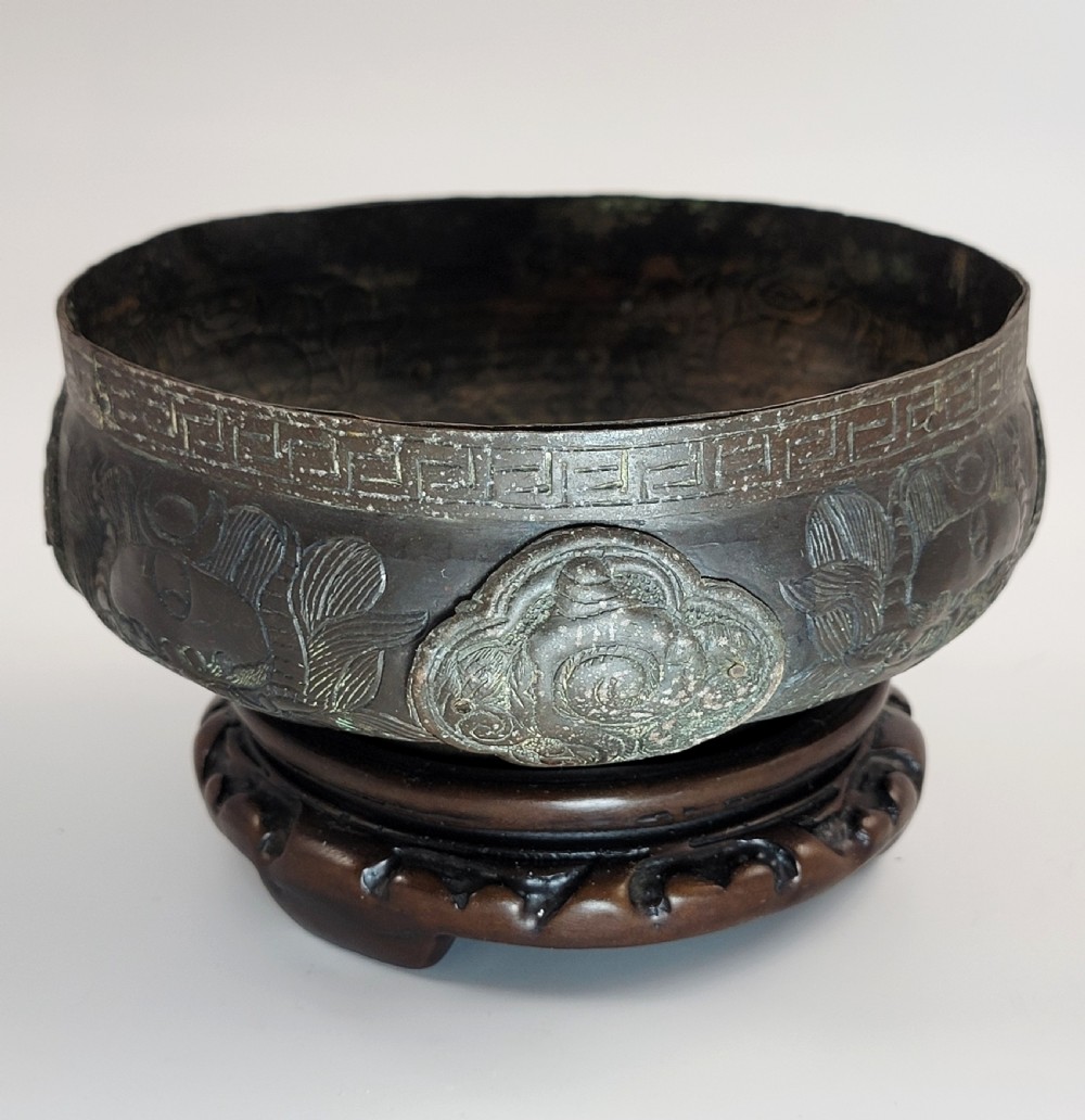 A patinated dark bronze Japanese bowl with carved archaic relief patterns and a key-fret border, resting on a carved wooden stand. View VII