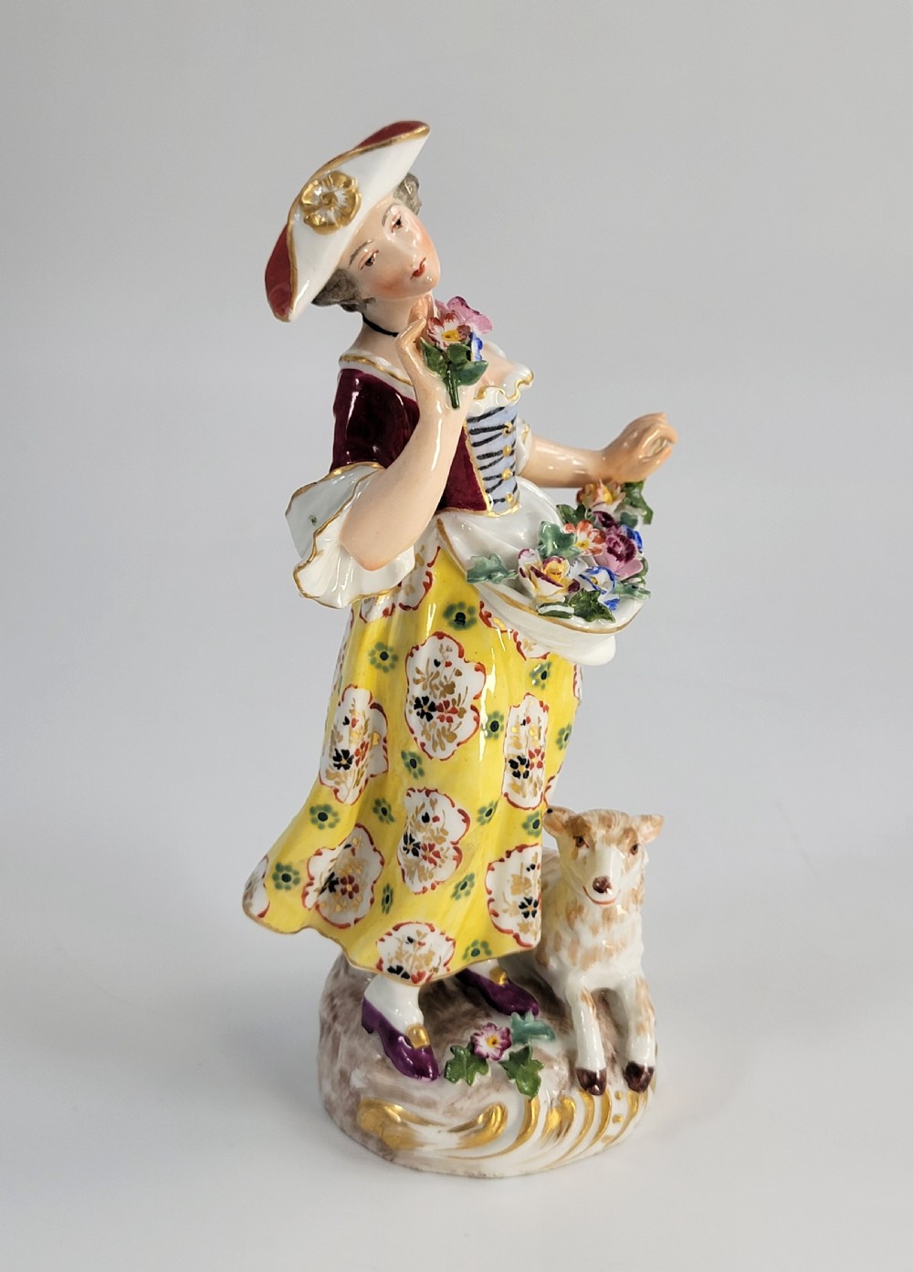 A 19th-century German porcelain figure depicting a shepherdess in a yellow patterned skirt with a lamb resting at her feet. View VI