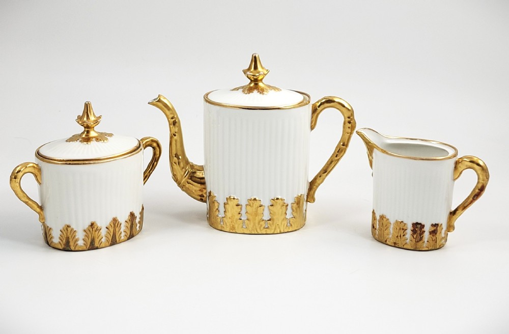 A three-piece white porcelain coffee set featuring a pot, sugar bowl, and cream jug with fluted bodies and gilded acanthus leaf detailing.