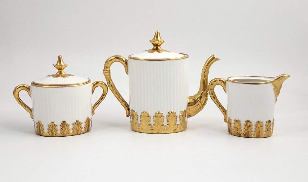 A three-piece white porcelain coffee set featuring a pot, sugar bowl, and cream jug with fluted bodies and gilded acanthus leaf detailing. View III