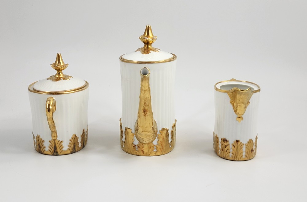 A three-piece white porcelain coffee set featuring a pot, sugar bowl, and cream jug with fluted bodies and gilded acanthus leaf detailing. View IV
