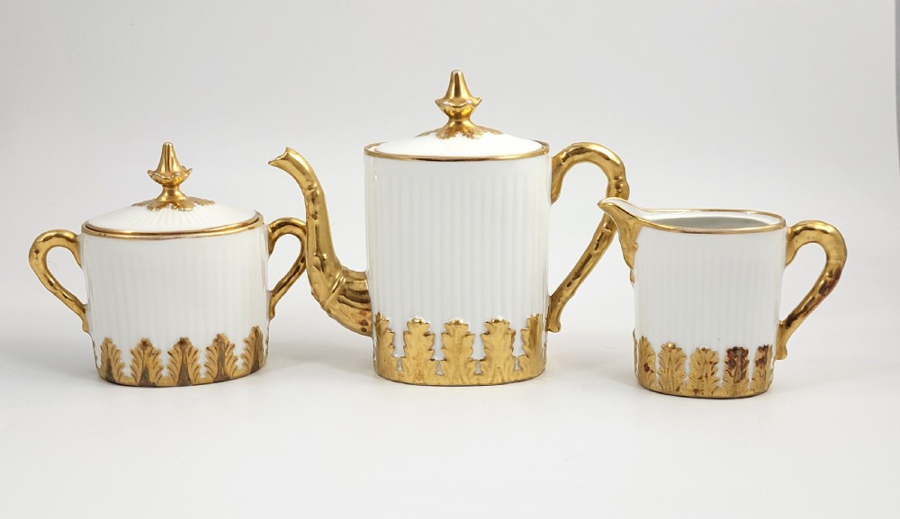A three-piece white porcelain coffee set featuring a pot, sugar bowl, and cream jug with fluted bodies and gilded acanthus leaf detailing. View V