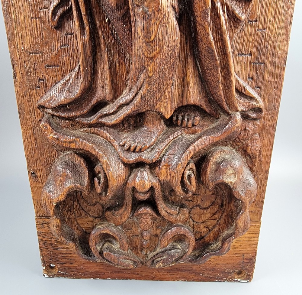A vertical carved oak panel displaying a high-relief figure of Faith holding a cross and a book beneath ornate Baroque scrollwork. View V