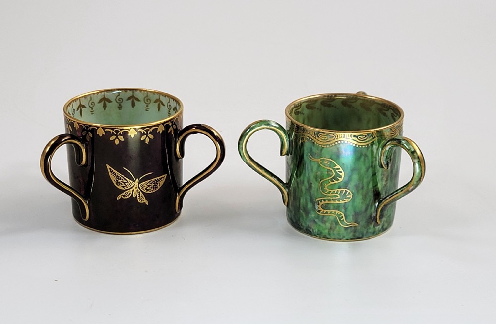 A pair of small three-handled ceramic cups, one in deep purple with a gilded butterfly design and the other in iridescent green with a gilded snake motif.
