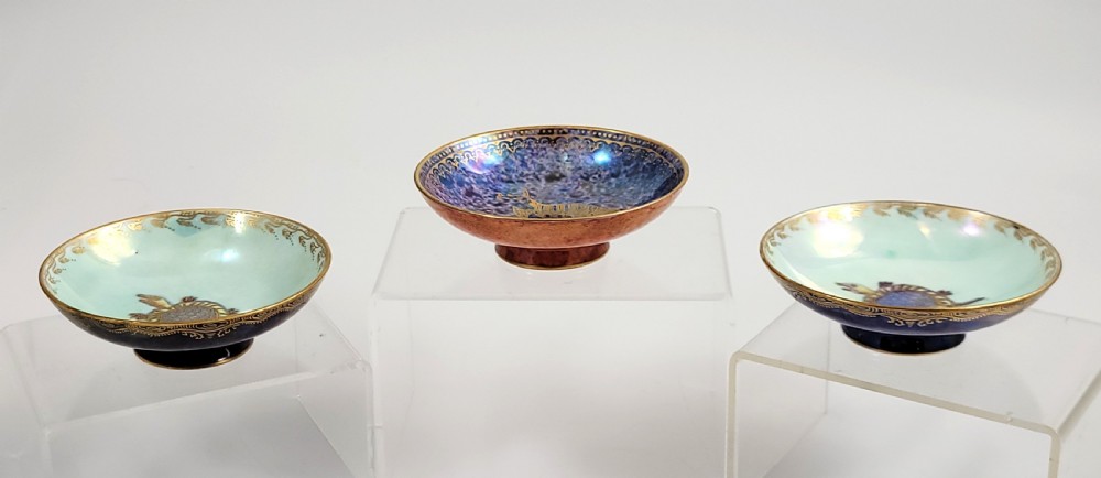 A set of three miniature footed porcelain bowls featuring iridescent lustre glazes in shades of blue, orange, and turquoise, embellished with gilded rim details and central motifs.