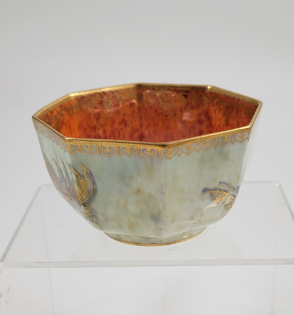 An octagonal ceramic bowl featuring a mottled pale blue and green iridescent exterior with gilt motifs and a bright orange mottled interior with gold borders.