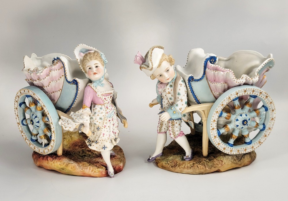 A pair of late 19th-century German hand-painted porcelain figural flower carts featuring a male and female figure in 18th-century attire pulling gilded two-wheeled carriages.