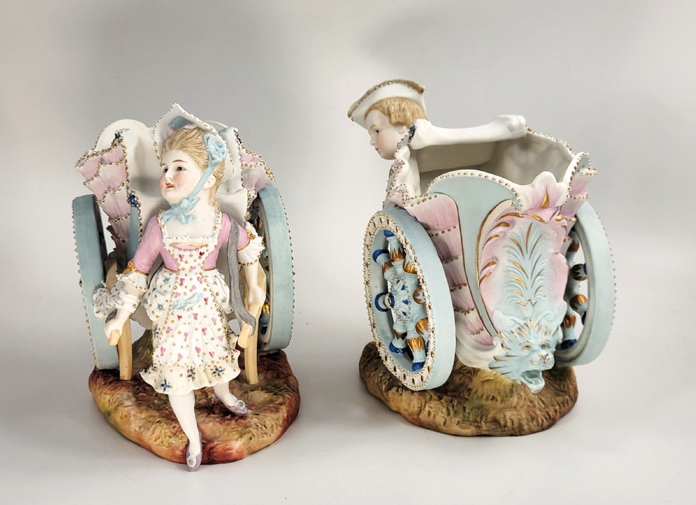 A pair of late 19th-century German hand-painted porcelain figural flower carts featuring a male and female figure in 18th-century attire pulling gilded two-wheeled carriages. View II