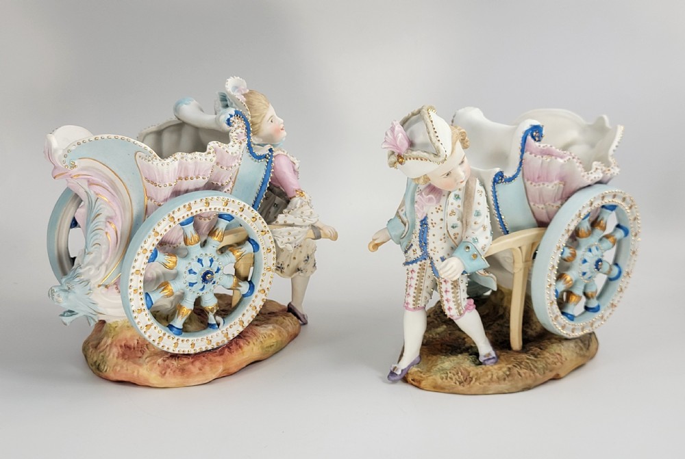 A pair of late 19th-century German hand-painted porcelain figural flower carts featuring a male and female figure in 18th-century attire pulling gilded two-wheeled carriages. View V