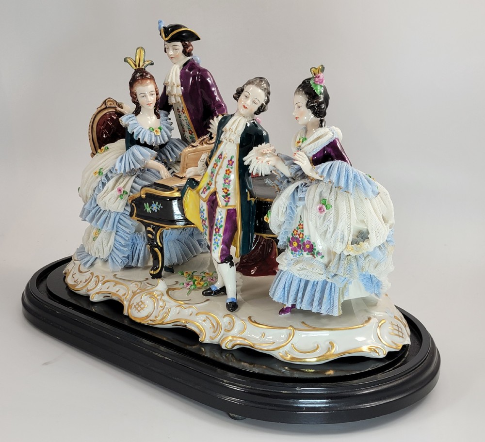 A German porcelain figural group depicting four 18th-century courtly figures around a grand piano with detailed porcelain lace dresses on a black wooden base. View II