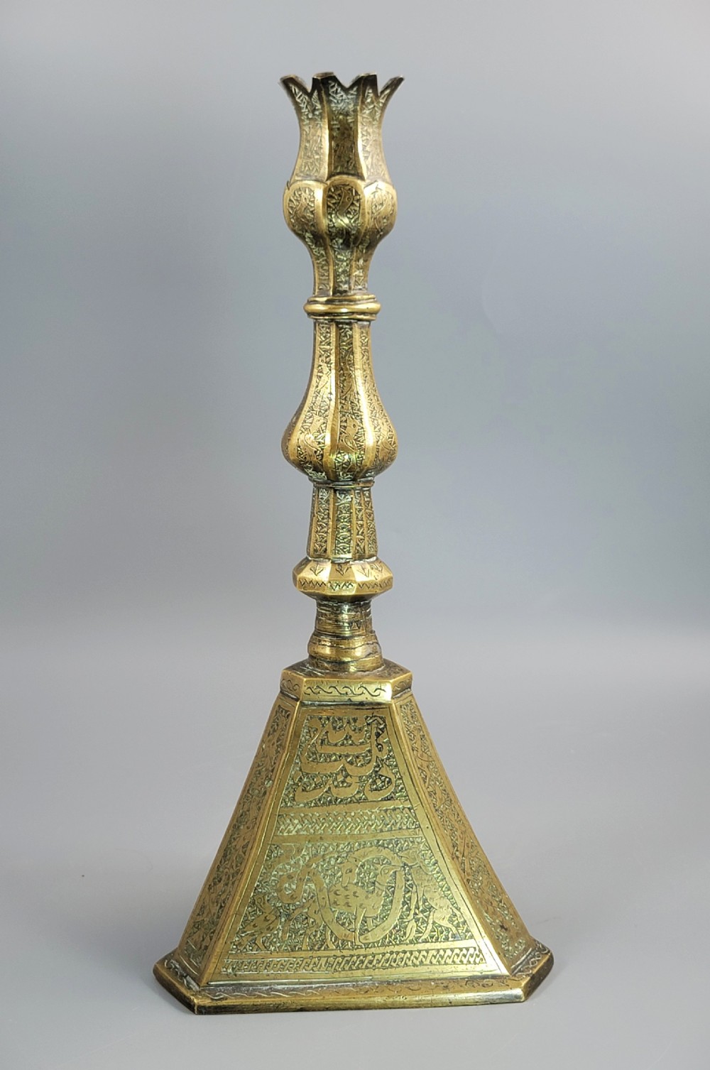 A tall late 19th-century Qajar Persian brass candlestick featuring a wide triangular engraved base, a knopped stem, and a flared lotus socket.