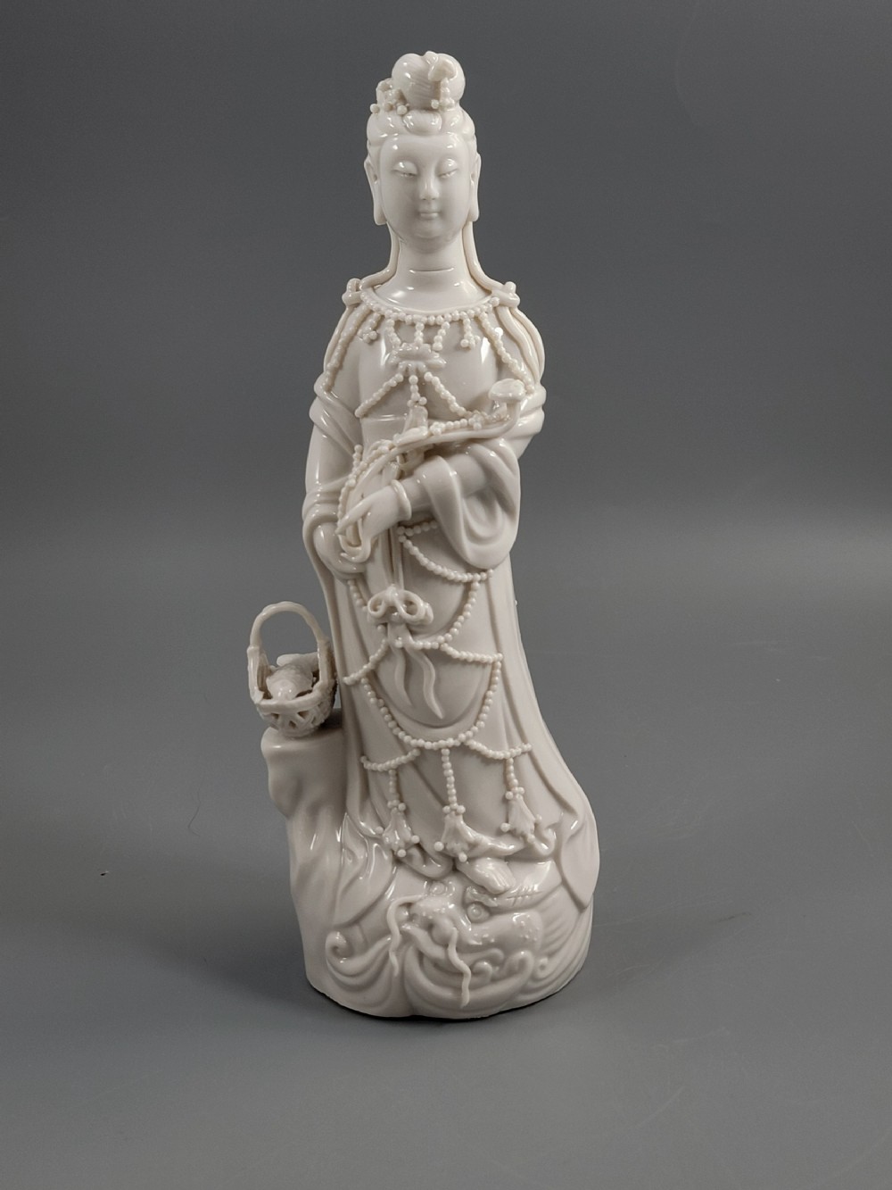 An early 20th-century Blanc de Chine porcelain figure of Guanyin wearing beaded robes and holding a fish basket on a wave pedestal.