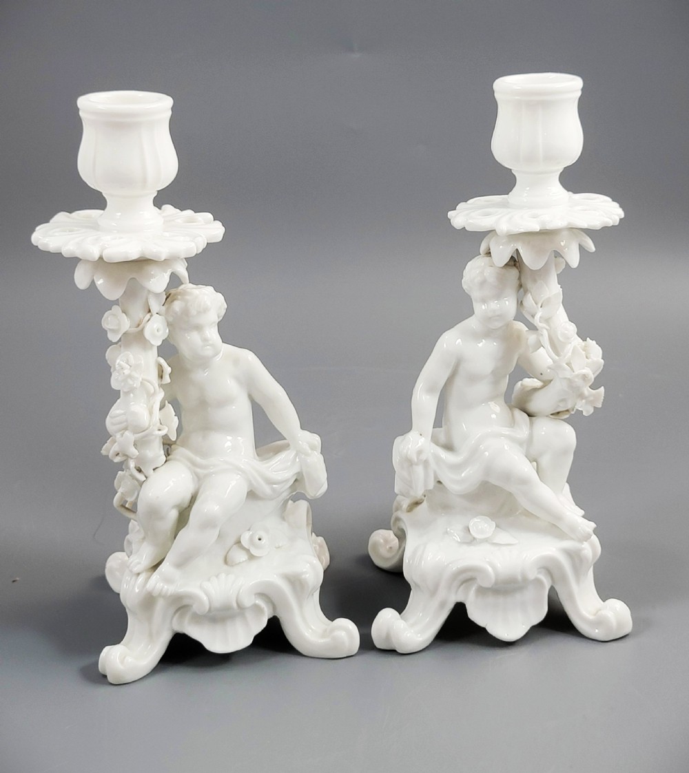 A pair of white glazed porcelain candlesticks featuring sculpted putti figures resting against floral encrusted columns on scrollwork bases.