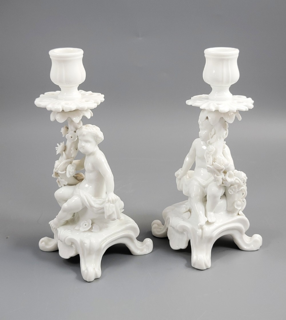 A pair of white glazed porcelain candlesticks featuring sculpted putti figures resting against floral encrusted columns on scrollwork bases. View II