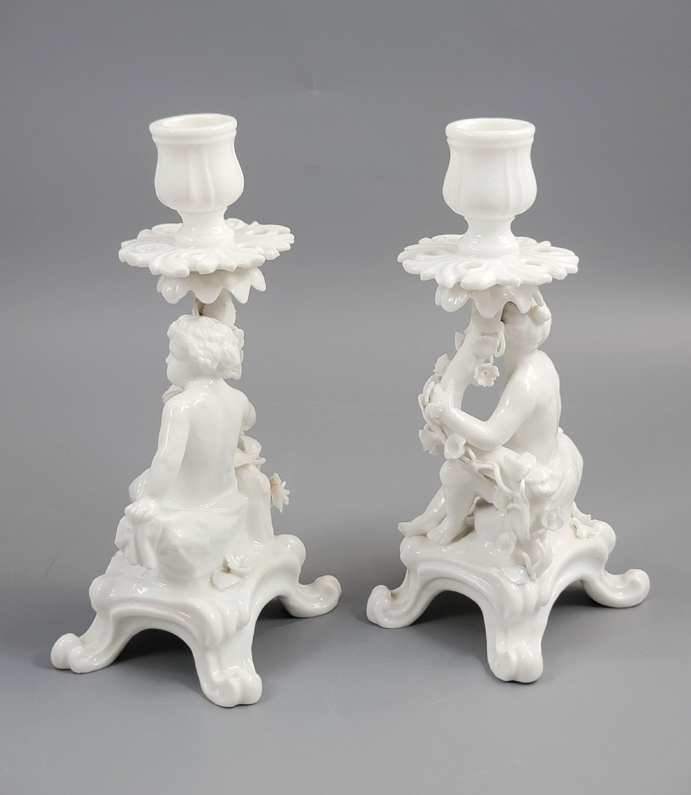 A pair of white glazed porcelain candlesticks featuring sculpted putti figures resting against floral encrusted columns on scrollwork bases. View III
