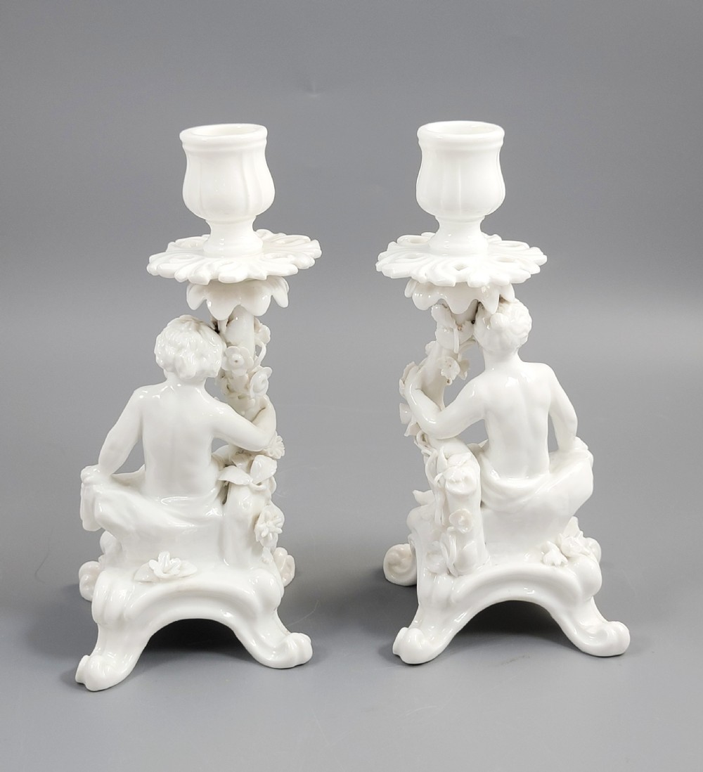 A pair of white glazed porcelain candlesticks featuring sculpted putti figures resting against floral encrusted columns on scrollwork bases. View IV