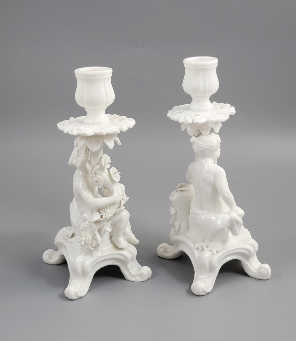 A pair of white glazed porcelain candlesticks featuring sculpted putti figures resting against floral encrusted columns on scrollwork bases. View V