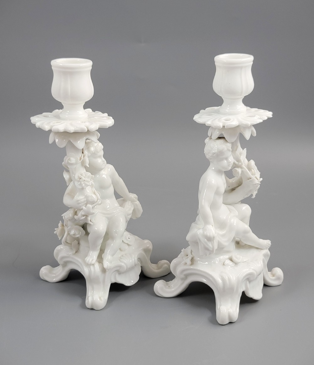 A pair of white glazed porcelain candlesticks featuring sculpted putti figures resting against floral encrusted columns on scrollwork bases. View VI