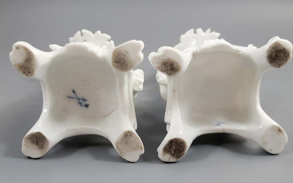 A pair of white glazed porcelain candlesticks featuring sculpted putti figures resting against floral encrusted columns on scrollwork bases. View VII