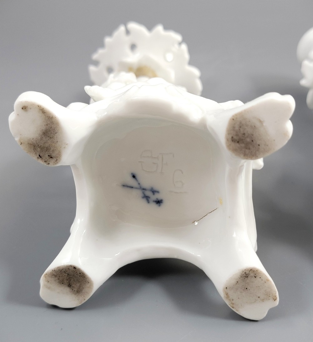 A pair of white glazed porcelain candlesticks featuring sculpted putti figures resting against floral encrusted columns on scrollwork bases. View VIII