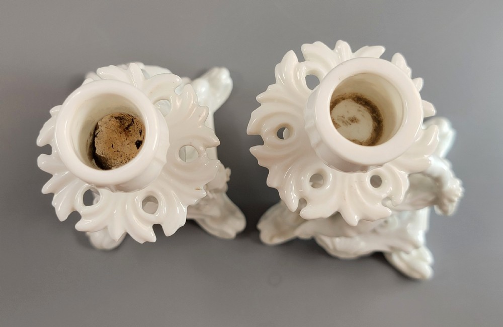 A pair of white glazed porcelain candlesticks featuring sculpted putti figures resting against floral encrusted columns on scrollwork bases. View X