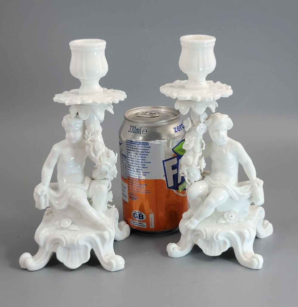 A pair of white glazed porcelain candlesticks featuring sculpted putti figures resting against floral encrusted columns on scrollwork bases. View XI