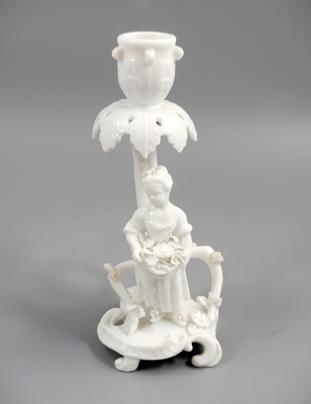 An antique white glazed porcelain candlestick featuring a moulded figurine of a girl holding flowers atop a Rococo scroll base.