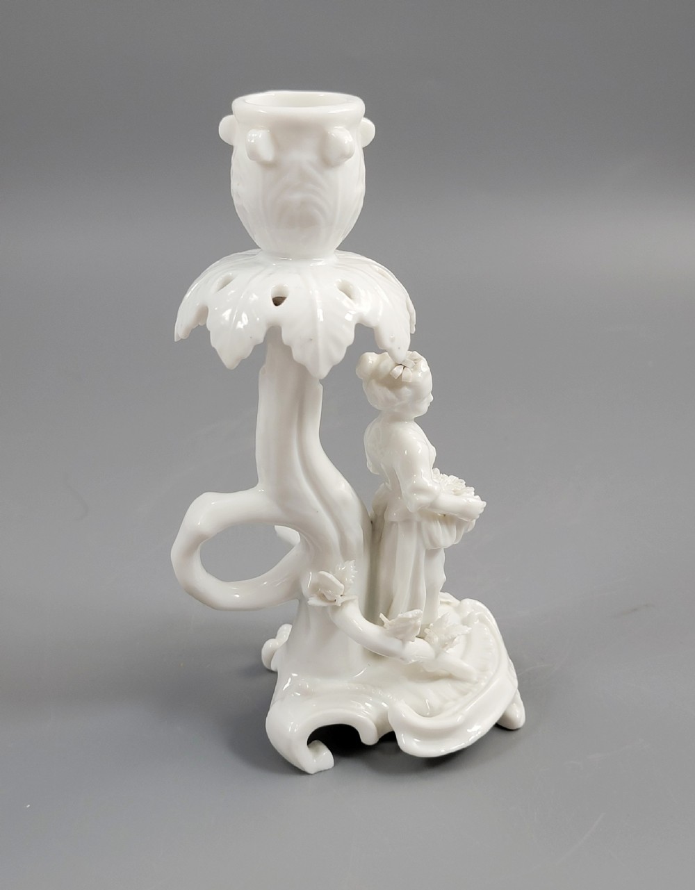 An antique white glazed porcelain candlestick featuring a moulded figurine of a girl holding flowers atop a Rococo scroll base. View V