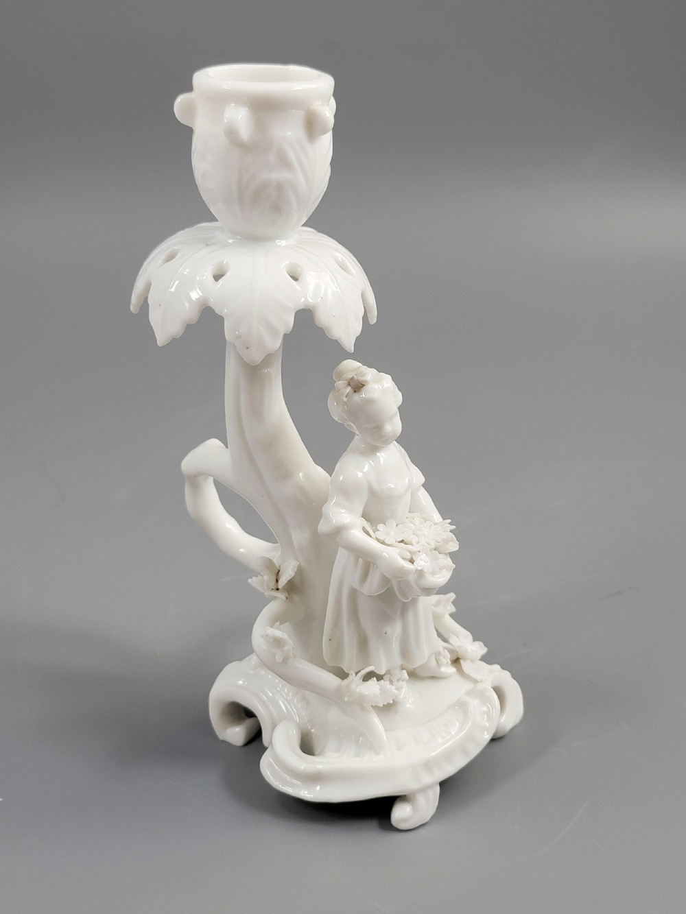 An antique white glazed porcelain candlestick featuring a moulded figurine of a girl holding flowers atop a Rococo scroll base. View VI