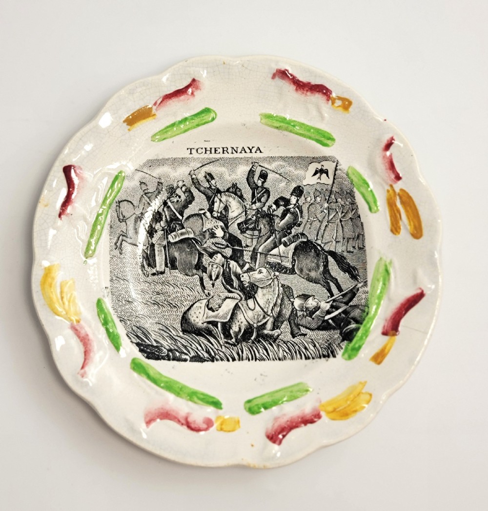 A circular Victorian ceramic plate featuring a central black transfer print of the Battle of Tchernaya surrounded by a scalloped rim with green, red, and yellow enamel accents.
