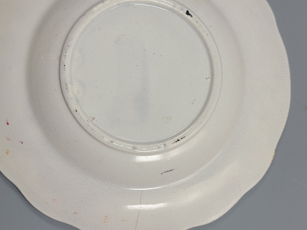 A circular Victorian ceramic plate featuring a central black transfer print of the Battle of Tchernaya surrounded by a scalloped rim with green, red, and yellow enamel accents. View V