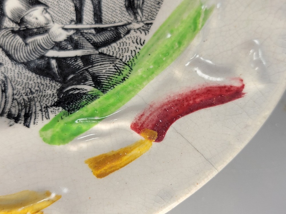 A circular Victorian ceramic plate featuring a central black transfer print of the Battle of Tchernaya surrounded by a scalloped rim with green, red, and yellow enamel accents. View VI