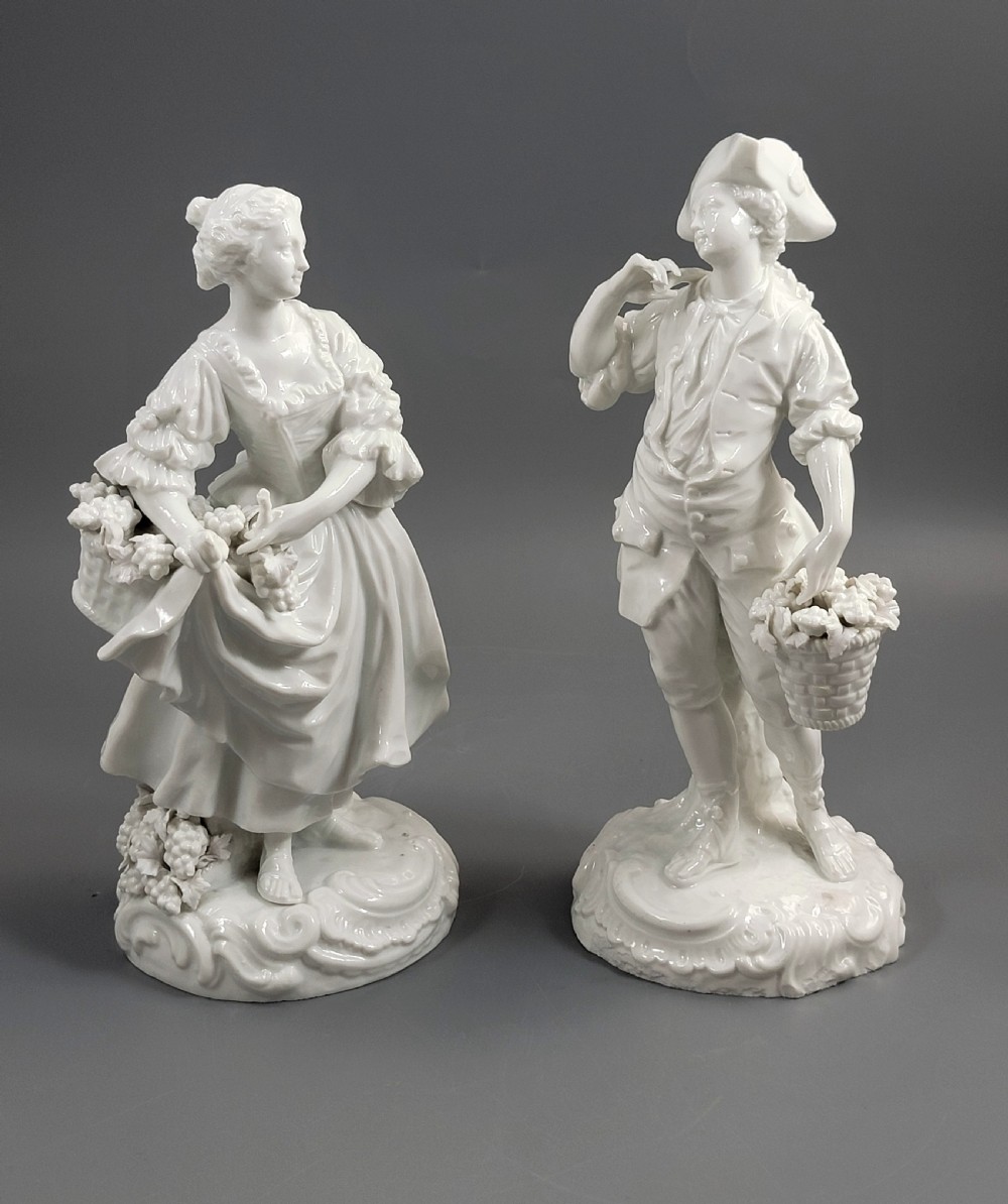 A pair of white glazed porcelain figurines depicting a man and woman dressed in 18th-century attire holding baskets filled with grapes on scrollwork bases.