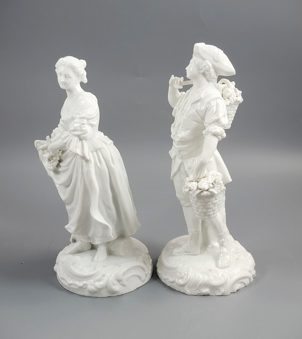A pair of white glazed porcelain figurines depicting a man and woman dressed in 18th-century attire holding baskets filled with grapes on scrollwork bases. View II