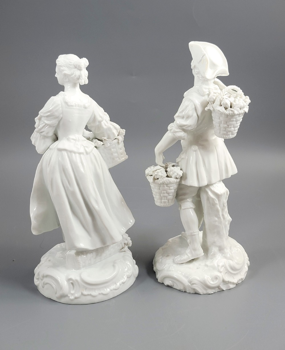 A pair of white glazed porcelain figurines depicting a man and woman dressed in 18th-century attire holding baskets filled with grapes on scrollwork bases. View III