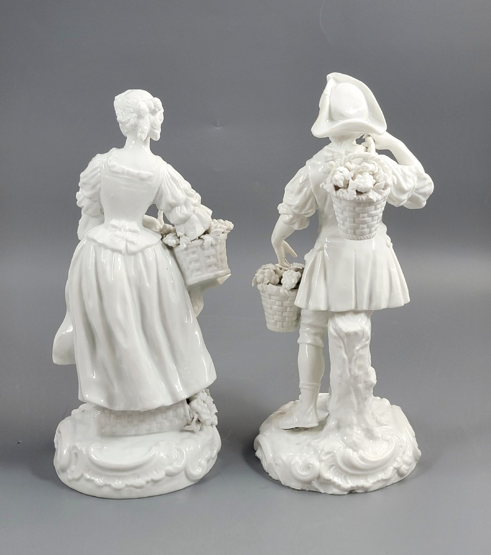 A pair of white glazed porcelain figurines depicting a man and woman dressed in 18th-century attire holding baskets filled with grapes on scrollwork bases. View IV