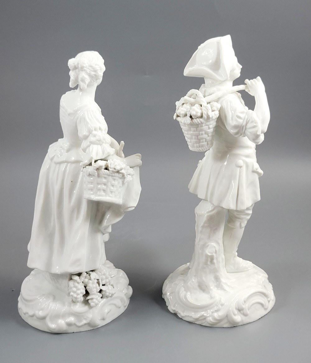 A pair of white glazed porcelain figurines depicting a man and woman dressed in 18th-century attire holding baskets filled with grapes on scrollwork bases. View V