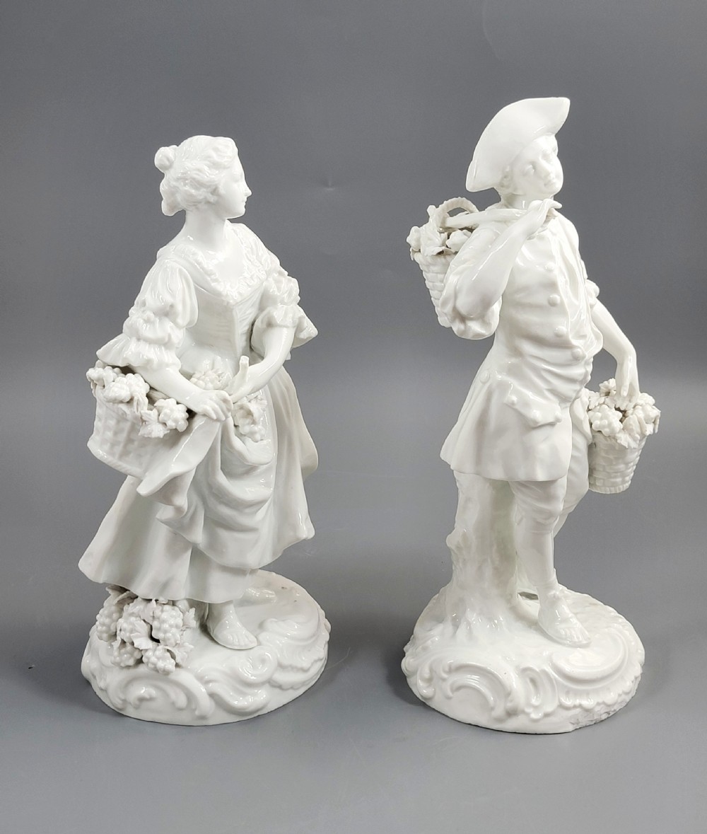 A pair of white glazed porcelain figurines depicting a man and woman dressed in 18th-century attire holding baskets filled with grapes on scrollwork bases. View VI
