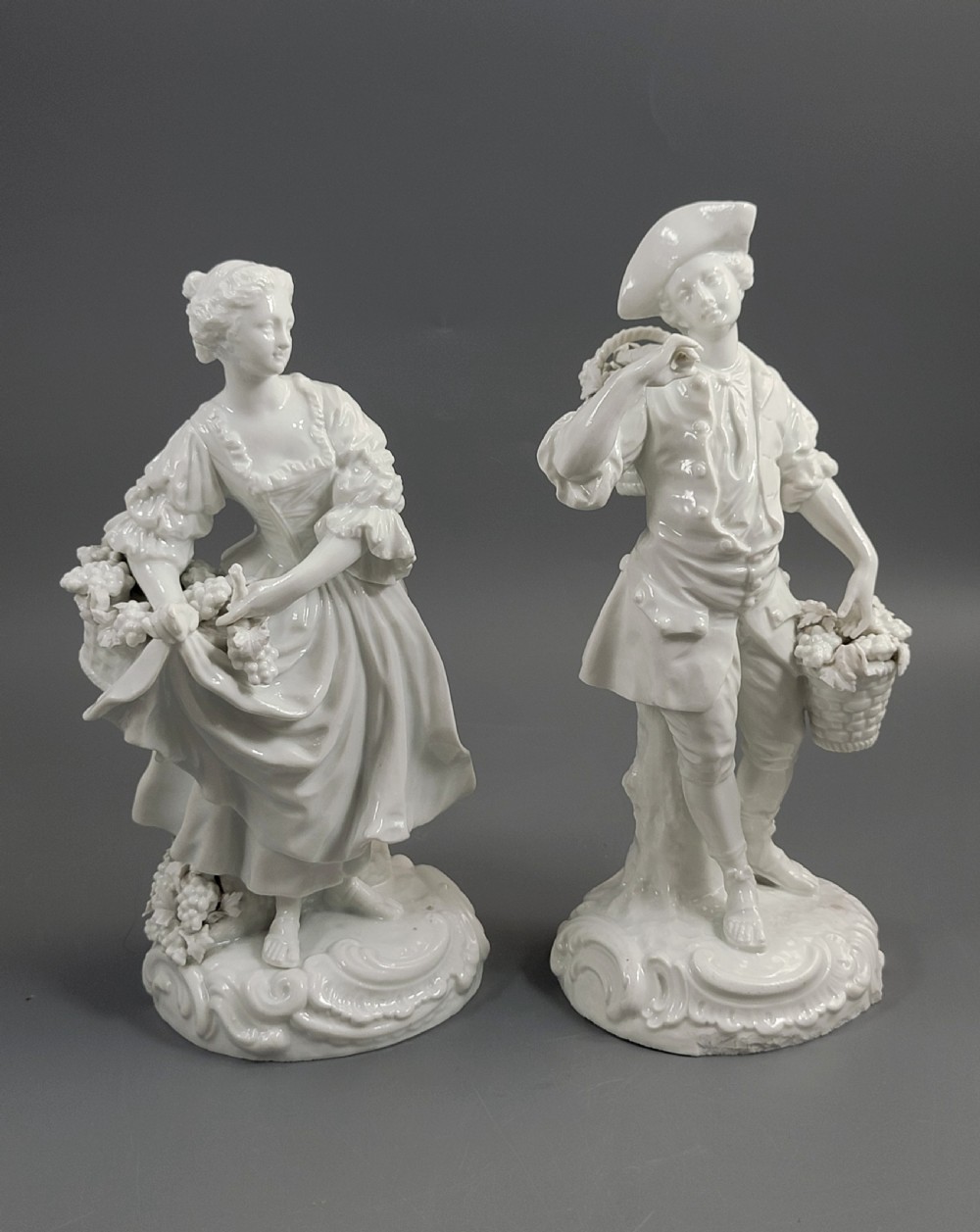 A pair of white glazed porcelain figurines depicting a man and woman dressed in 18th-century attire holding baskets filled with grapes on scrollwork bases. View VII