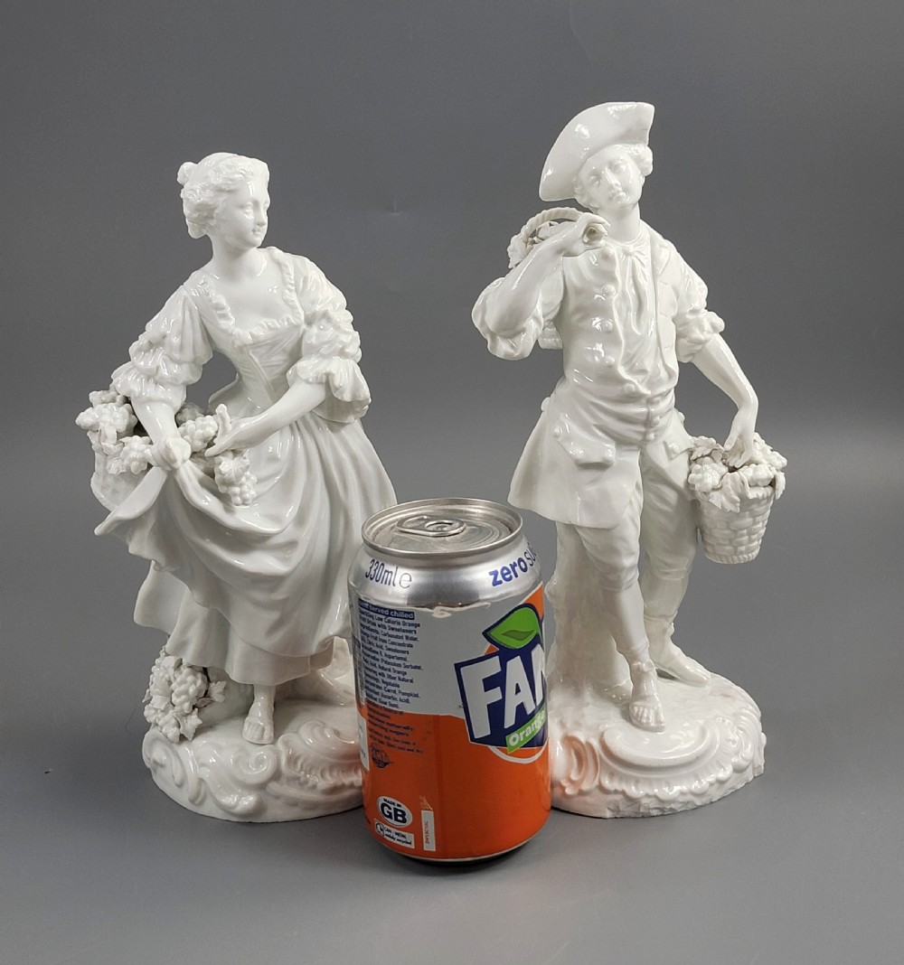 A pair of white glazed porcelain figurines depicting a man and woman dressed in 18th-century attire holding baskets filled with grapes on scrollwork bases. View VIII