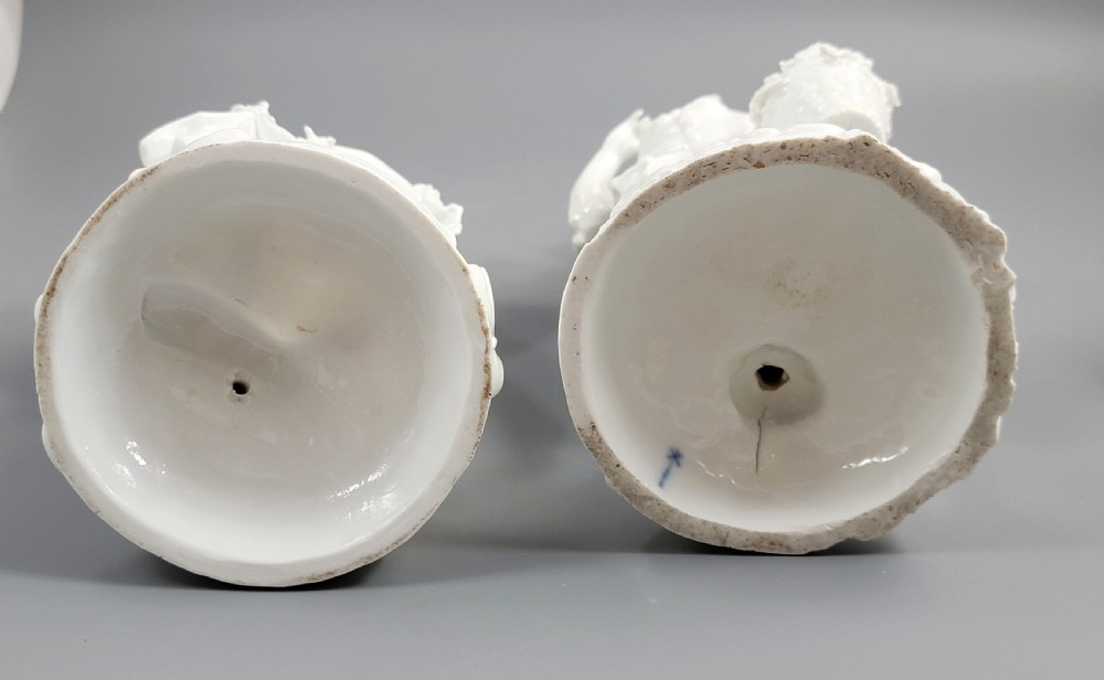 A pair of white glazed porcelain figurines depicting a man and woman dressed in 18th-century attire holding baskets filled with grapes on scrollwork bases. View IX