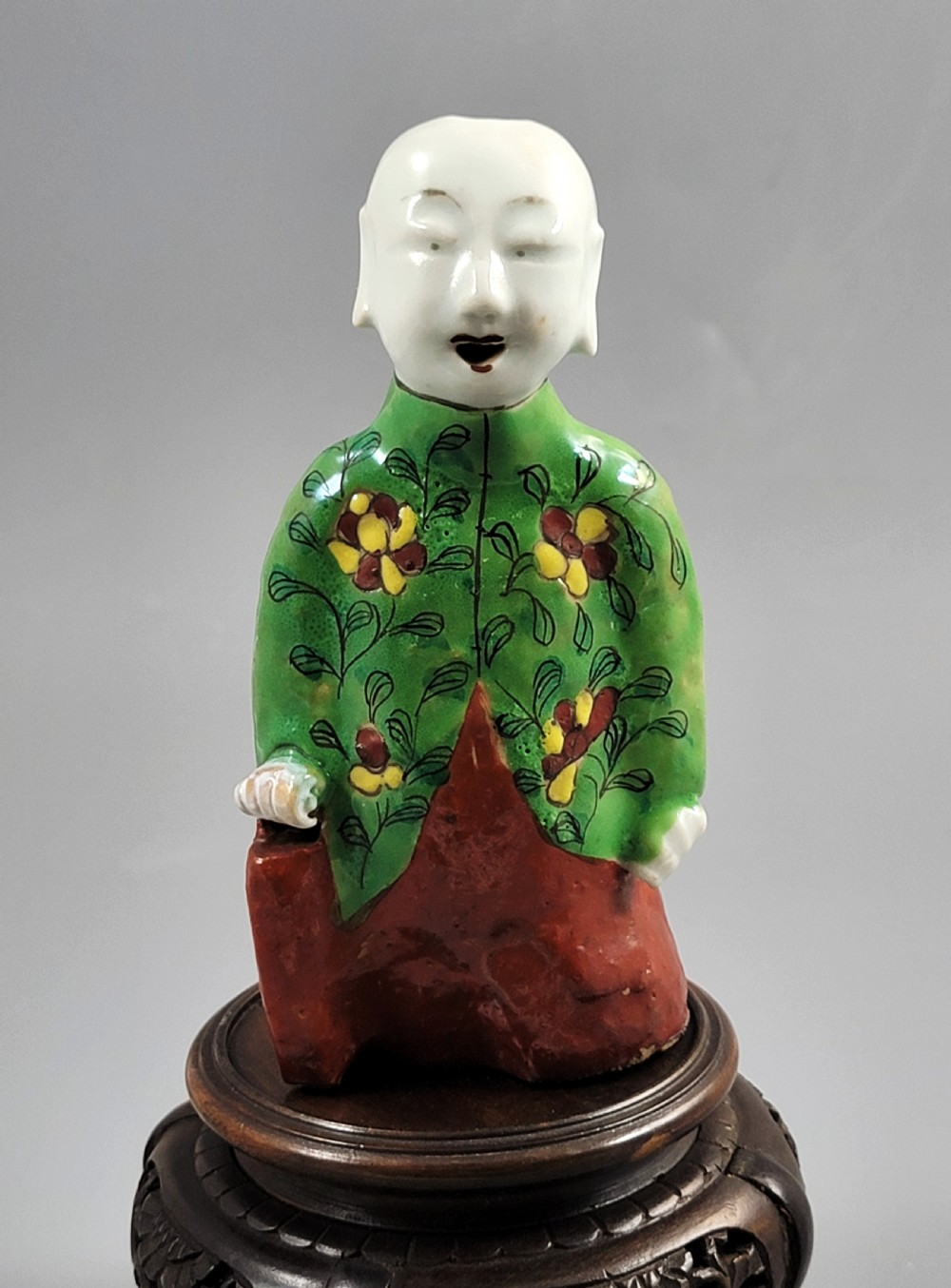 An antique Chinese ceramic incense holder depicting a smiling boy in a green floral jacket seated on a red base.