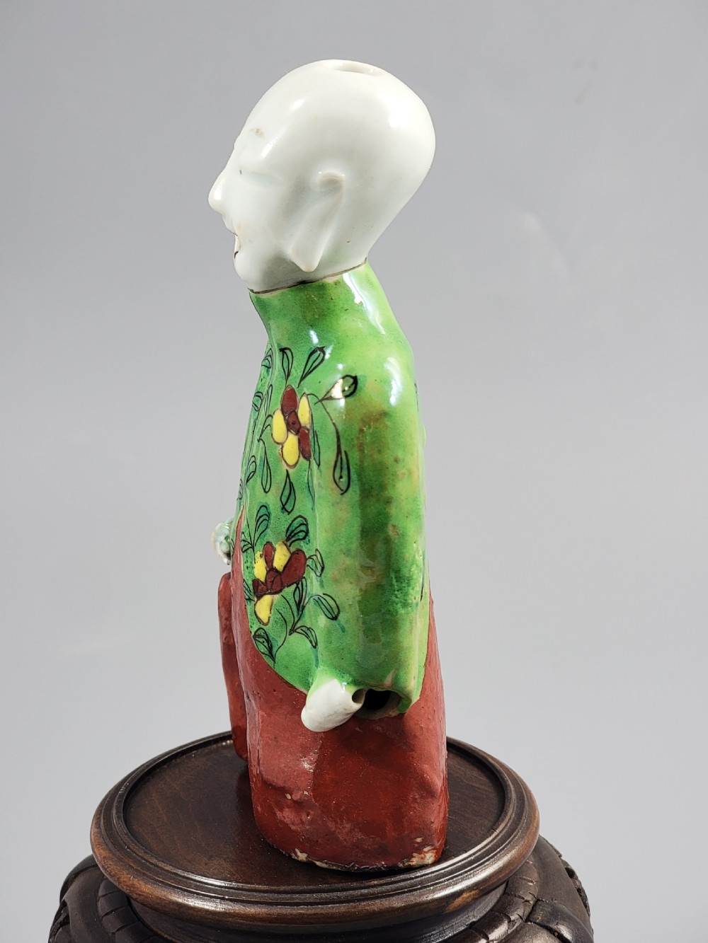 An antique Chinese ceramic incense holder depicting a smiling boy in a green floral jacket seated on a red base. View II